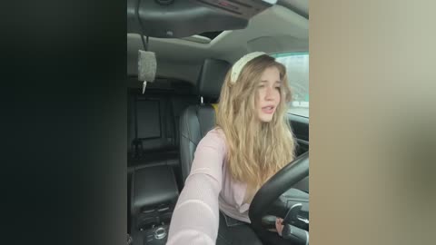 A video captures a young woman with long, wavy blonde hair, wearing a pink cardigan, driving a car. The interior is modern, with black leather seats and a beige dashboard.