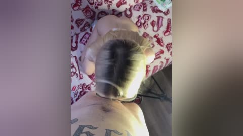 Media: Video of a blonde woman, lying face down on a red and white patterned bedspread, showing her back and shoulder tattoos.