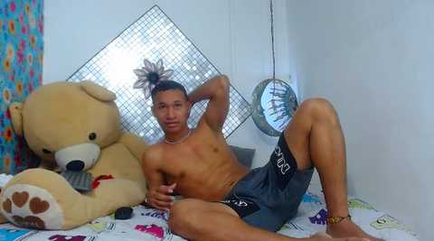 Video of a shirtless, muscular young man with light brown skin and short dark hair, wearing gray shorts, reclining on a bed with a large stuffed bear and a mirror behind him.