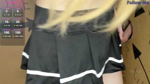 Media: Video of a person with long blonde hair, wearing a black skirt with white stripes, holding a phone. Background shows a poster with stats and the \"FollowMe\" logo.