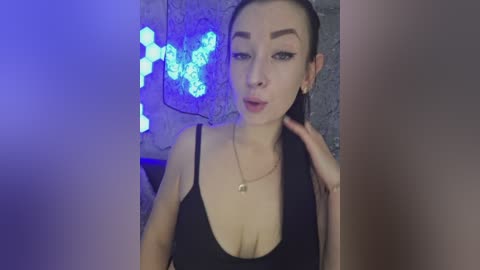 A video of a young woman with fair skin, dark hair pulled back, wearing a black tank top, with a tattoo on her neck. She stands against a blue hexagonal light backdrop, indoors.