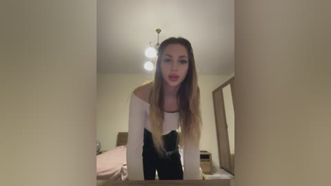 Media: A video of a blonde woman in a white off-the-shoulder top and black pants, leaning over a mirror, with a beige room and a wooden door in the background.