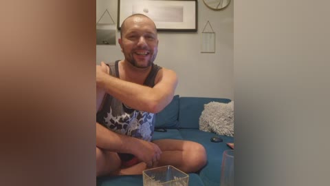 Video of a muscular, bald man with a beard in a gray tank top, sitting on a blue couch, smiling, holding a glass, in a modern living room with minimalist decor.