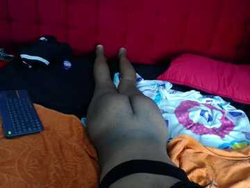 Video of a person lying on a bed with a red headboard, wearing black tights and black underwear, with a colorful blanket and orange sheets, and a keyboard nearby.