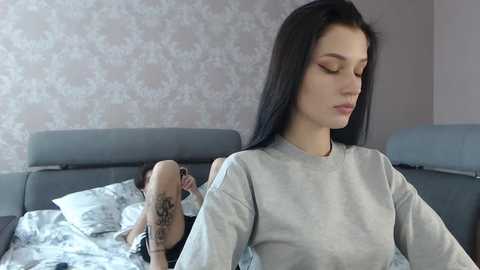 Media: Video of a young woman with long black hair, fair skin, and a tattoo on her arm, sitting on a bed in a gray shirt, looking serene. Background features a gray headboard and floral-patterned wallpaper.