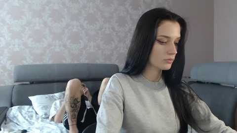 Media: Video of a woman with long black hair in a gray sweater, sitting on a bed with floral wallpaper and a black-haired man lying behind her.