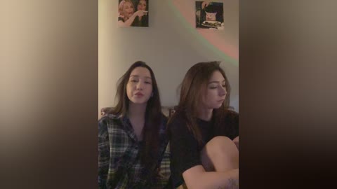 Media: Video of two young women with long hair, one in a plaid shirt and the other in a black top, seated side-by-side, with a rainbow reflection on the wall.