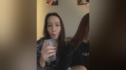 Media: A video of a young woman with long dark hair, wearing a plaid shirt, sipping from a glass while another person, with shoulder-length red hair, is seated behind her.