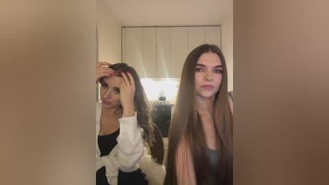 Media: Video of two young women in a narrow hallway. One with long brown hair, black top, and white jacket, touches her hair; the other, with long straight hair, wears a dark top.