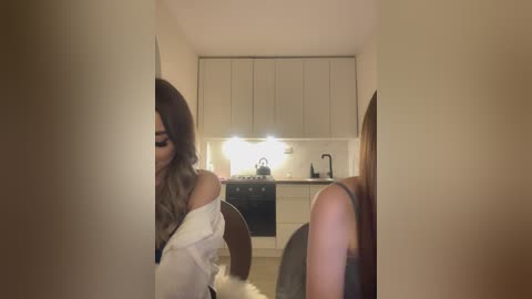 Media: Video of a dimly lit, narrow hallway with beige walls. A woman with long, wavy brown hair, wearing a white off-shoulder blouse, is partially visible in the left foreground. The background features a modern kitchen with white cabinets, a black stove, and a black kettle.