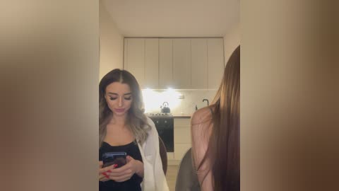 Media: A video of a young woman with light skin and long, wavy, brown hair, wearing a black dress and white cardigan, taking a selfie in a modern, minimalist kitchen with white cabinets and stainless steel appliances.