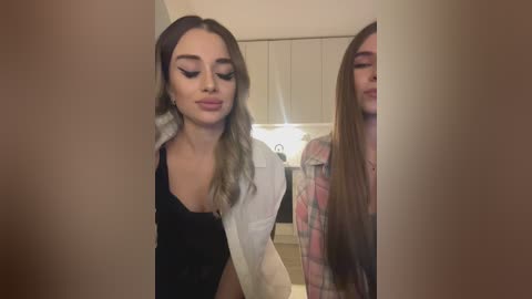 Media: Video of two young women with long, straight hair, one with light brown hair, the other with dark brown hair, standing side by side in a modern kitchen with white cabinets and a sink. They are wearing casual clothes and smiling.