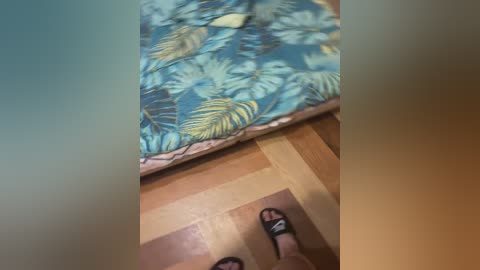 A video showing a person's legs in black sandals on a wooden floor, with a vibrant, tropical-patterned bedspread in the background. The image has a soft, blurred effect.