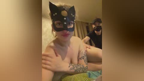 Video of a fair-skinned woman with large breasts and tattoos, wearing a black cat mask with rhinestones, sitting topless on a bed with a man in the background, both in a dimly lit room.