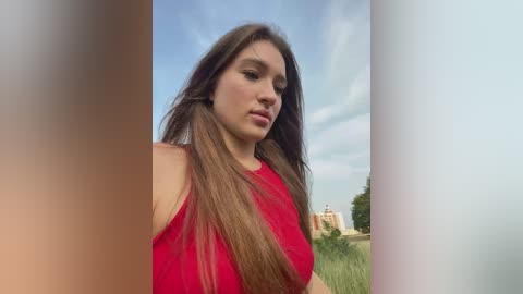 Media: Video of a young Caucasian woman with long, straight brown hair, wearing a sleeveless red dress, standing outdoors against a cloudy sky and grassy field.