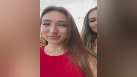 Media: Video of two young women with light skin and straight, long brown hair, wearing red tops. The background is blurred, with a hint of a white ceiling.