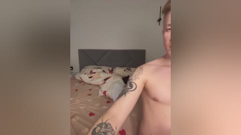 Media: Video of a nude, fair-skinned man with tattoos on his left arm and torso, seated on a bed with patterned sheets, in a dimly lit bedroom.