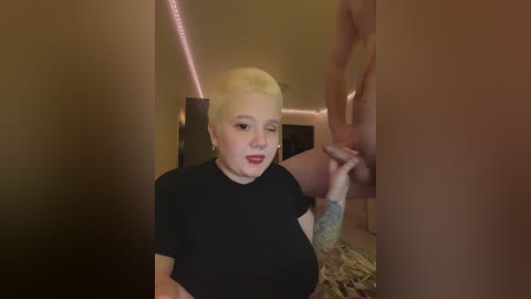 Video of a short-haired, pale-skinned woman with red lipstick, wearing a black top, holding a man's erect penis. Background features a dimly lit, beige room with a bed.