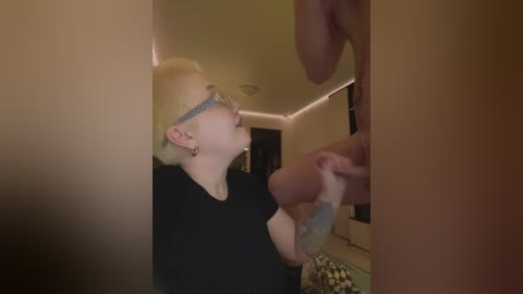 Video of a fair-skinned, blonde woman with glasses and tattoos, kneeling and performing oral sex on a man in a dimly lit room.