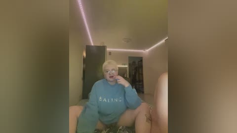 A video of a plus-size, fair-skinned woman with short blonde hair, wearing a blue Balenciaga sweatshirt and shorts, sitting on the floor in a dimly lit room with pink LED lights on the ceiling.
