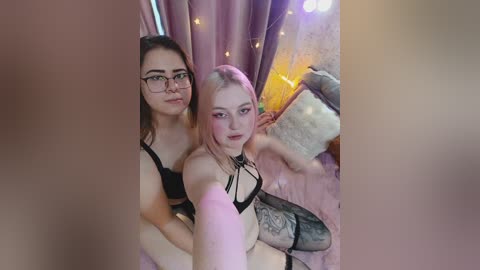 A video features two young women, one with glasses and light skin, the other with pink hair, wearing black lingerie, taking a selfie in a cozy, softly lit room with pink curtains.