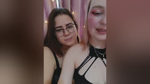Video of two young women with fair skin, one wearing glasses and black lingerie, the other with light makeup, standing in front of pink curtains.