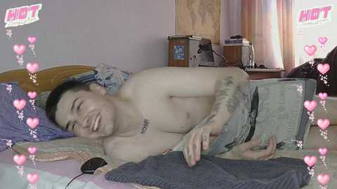 A video of a shirtless Asian man with tattoos, lying on a bed in a dimly lit bedroom, laughing, with heart-shaped filters overlaying the image.