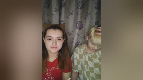 Media: Video of two young women in a cozy, dimly lit room. One wears a red shirt with text, the other a checkered shirt. Both have long hair. Floral curtains in the background.