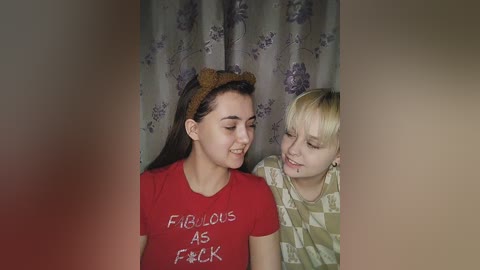 Media: Video of two young girls, one with dark hair and the other blonde, smiling in front of floral curtains. They wear casual t-shirts.