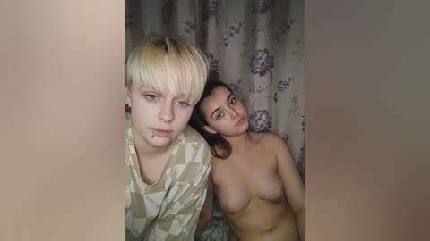 Media: Video of two young women, one with short, blonde hair and another with long, dark hair, both topless, leaning against floral-patterned curtains in a dimly lit room.