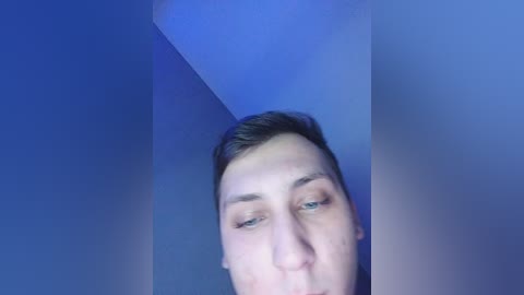 Video of a young man with short brown hair, blue eyes, and pale skin, captured from a low angle, against a blue-painted wall.