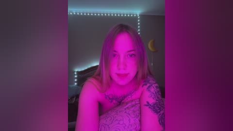 Media: Video of a smiling, light-skinned woman with blonde hair, sitting in a dimly lit bedroom. She has tattoos on her arms and shoulders, and the room has a purple glow from string lights.