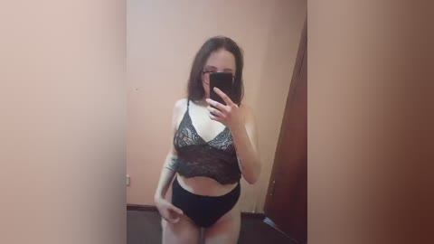 Video of a slender, fair-skinned woman with shoulder-length brown hair, wearing a black lace bra and high-waisted panties, taking a mirror selfie in a dimly lit room with peach-colored walls.