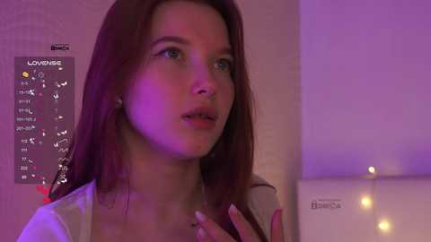 Video of a young woman with fair skin and long, straight brown hair, wearing a white top, illuminated by purple and pink lighting. Background shows a white wall and a light fixture with small, warm bulbs.