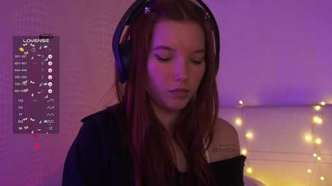 Video of a young woman with long auburn hair, wearing black headphones, eyes closed, in a softly lit room with purple and yellow lights.