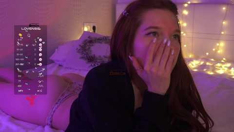 A video of a young woman with long, red hair, wearing a black top, covering her mouth, and lying on a bed with white sheets and fairy lights.