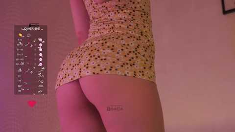 Video of a woman with fair skin, wearing a short, patterned dress, highlighting her round buttocks. Background shows a calendar with dates and a blurred wall.