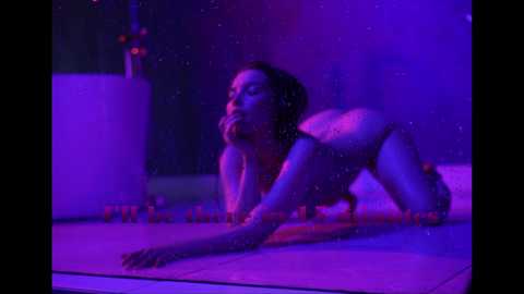 A dimly lit, grainy video of a nude woman with fair skin and shoulder-length dark hair, posed seductively on all fours, bathed in purple and pink hues.