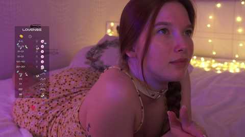 Video of a young Caucasian woman with light skin and long brown hair in a braid, wearing a floral dress, lying on a bed with white sheets. The room is softly lit with warm, glowing fairy lights.