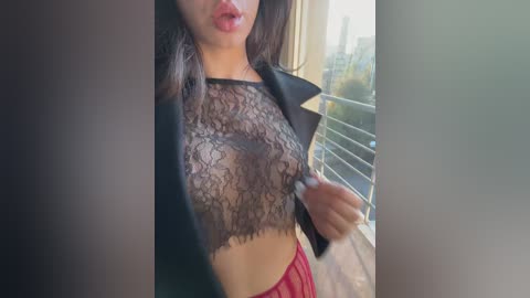 Media: Video of a woman with medium brown skin, wearing a sheer black lace crop top revealing her nipples, red panties, and a black leather jacket. She stands indoors near a window with a blurred cityscape in the background.