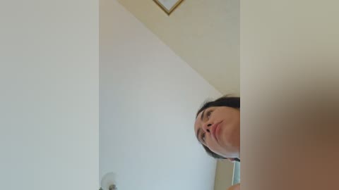 Media: A video of a young woman with light skin and dark hair, partially obscured by a white ceiling corner, looking down with a contemplative expression. The background features a beige wall and a small framed picture.