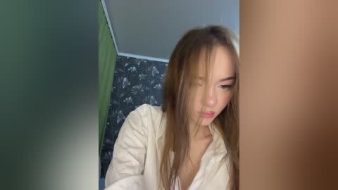 Media: A video captures an Asian woman with long brown hair, wearing a white shirt, looking down in a room with floral-patterned wallpaper and a green wall.