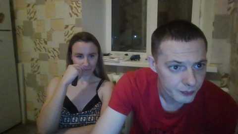 Video of a young woman with shoulder-length hair in a black top, and a young man with short hair in a red shirt, both looking at the camera. Background includes a window and tiled wall.