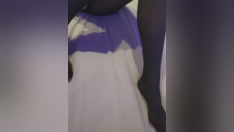 Video of a woman's upper body, wearing a white dress with a purple sash, seen from a low angle. The background is blurred, focusing on the dress's fabric and sash details.