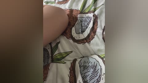 Video of a person lying on a bed with a white duvet featuring cartoonish coconuts and green palm leaves. The person's hand is partially visible, indicating a relaxed, casual pose.