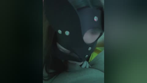 A blurry, dark video shows a person in a black, mask-like headpiece with reflective spots, kneeling on a bed with a yellow blanket. The scene is dimly lit, creating a mysterious atmosphere.