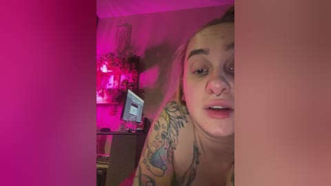A video of a young woman with light skin, long blonde hair, and tattoos, wearing a tank top, in a dimly lit room with pink lighting. She has a septum piercing and appears to be looking at something off-camera.