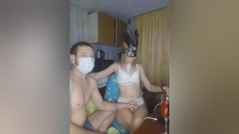 Media: Video of a young man and woman in a dimly-lit, cluttered room. Both are shirtless and wearing masks, sitting on a bed with a colorful blanket. The woman has bunny ears.