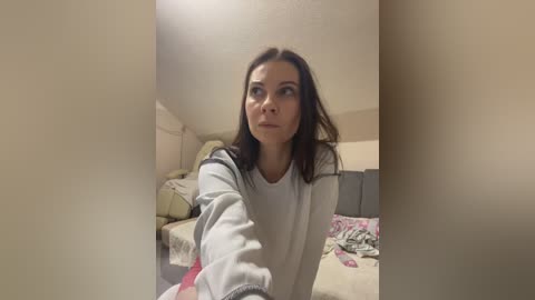 Media: Video of a young woman with dark hair and light skin, wearing a gray sweater, standing in a dimly lit bedroom with messy bedding.