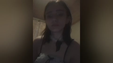 A blurry, dimly-lit video of a young woman with light skin and shoulder-length brown hair, wearing a black lace camisole and white ruffled collar, standing in a dimly-lit room with beige walls and a window with sheer curtains.